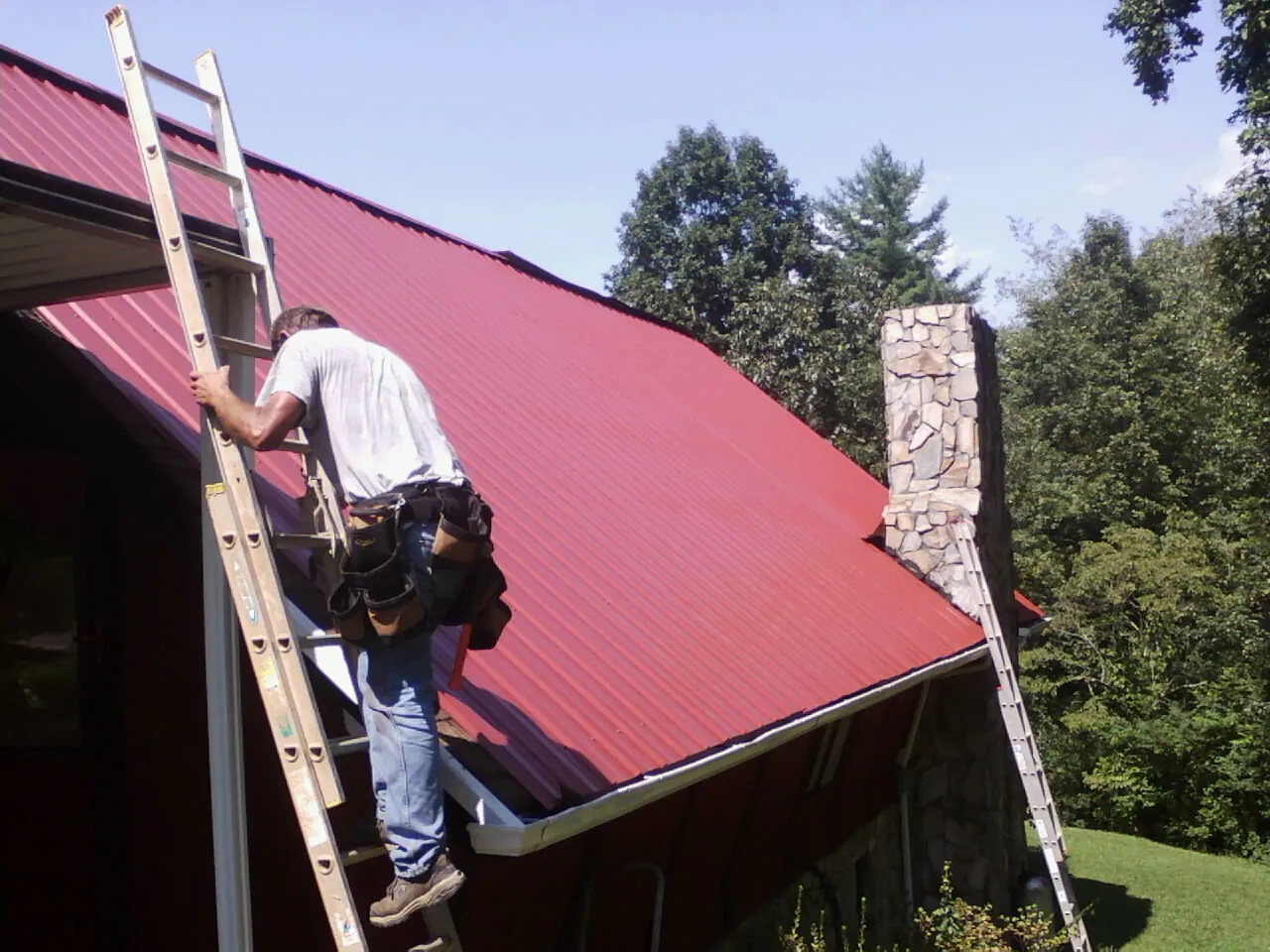 Roofer on a ladder beside a red metal roof and stone chimney — Soffit & Fascia Repair by Roofing Brothers in Newfoundland